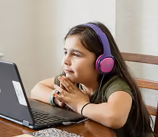 Young girl with long dark hair wearing pink and purple headphones, resting her chin on her hands and looking thoughtfully to the side while sitting at a table with a laptop.